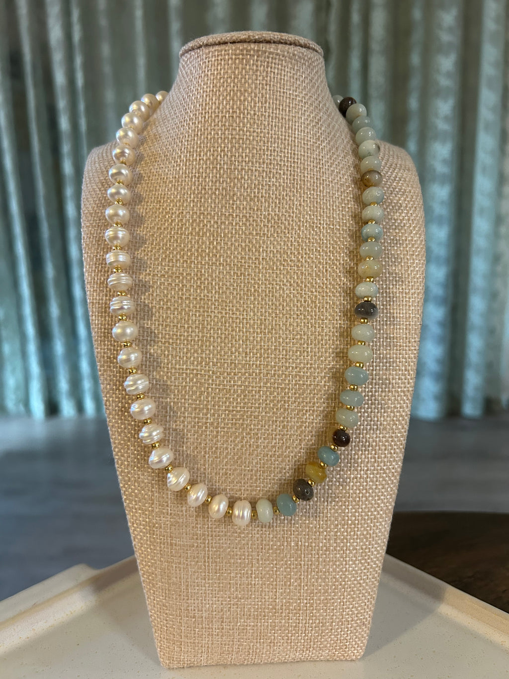 Beaded blue and pearl necklace on a beige bust with a green curtain background.