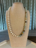 Beaded blue and pearl necklace on a beige bust with a green curtain background.