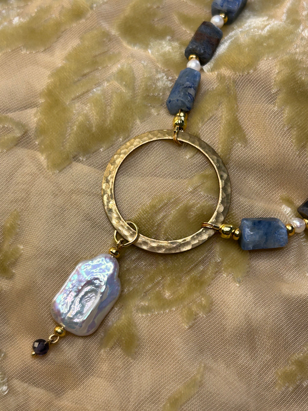Necklace with gold hoop, blue beads, and a white stone on a beige fabric background