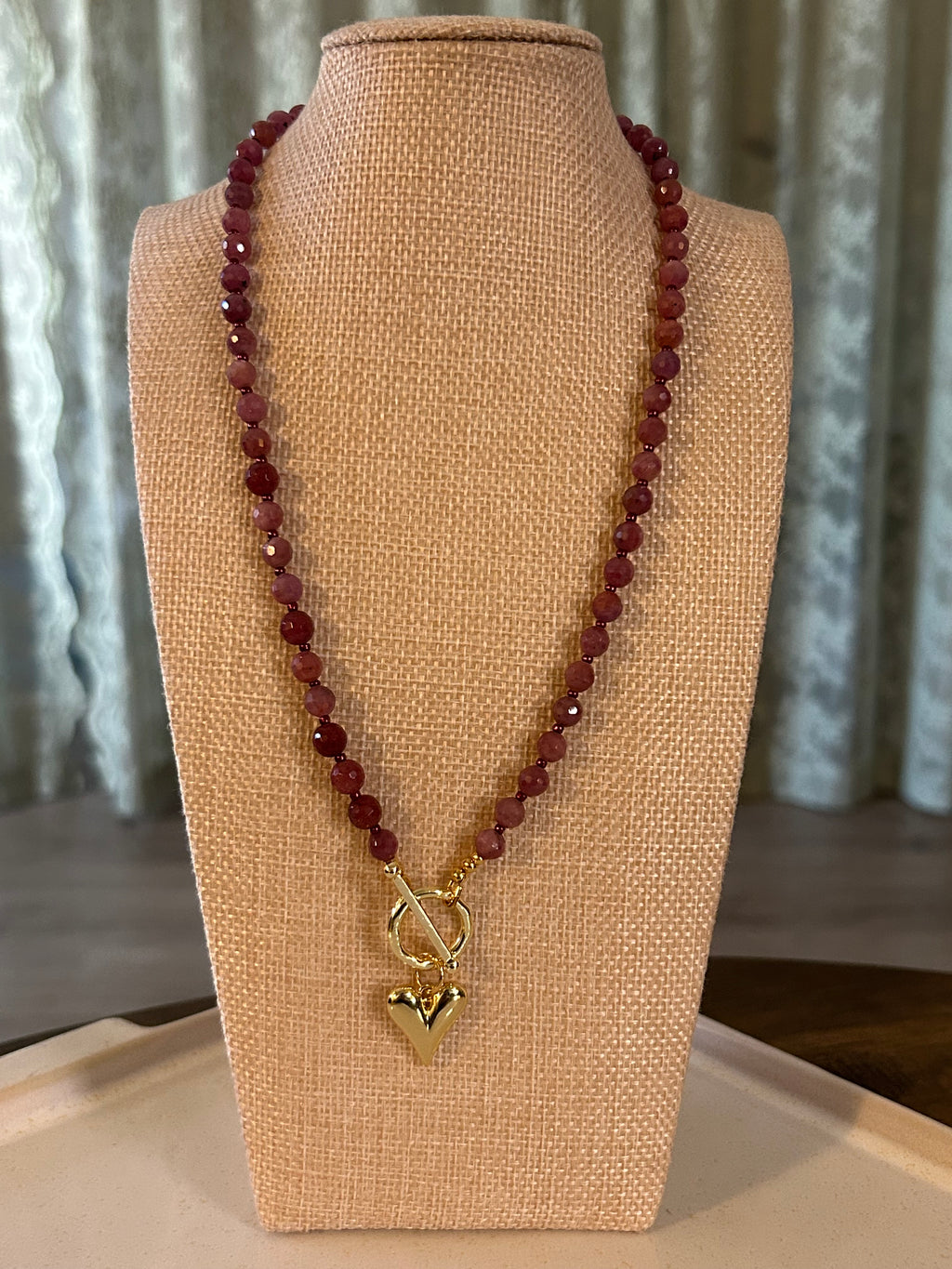 Necklace with red beads and gold clasp on a beige display stand.