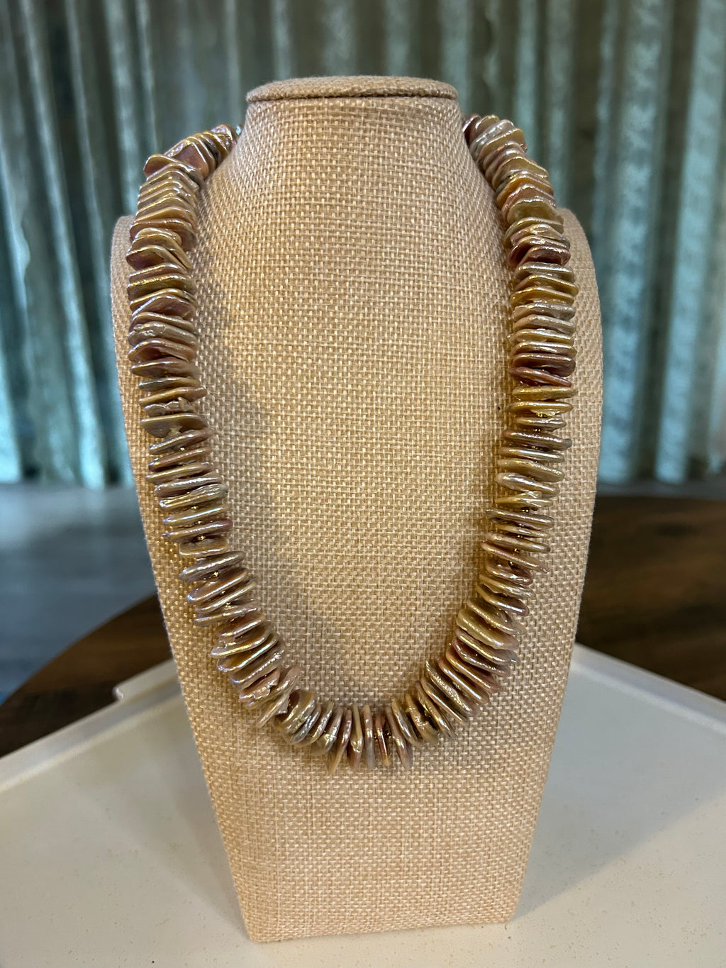 Beige necklace holder displaying a shell rondelle necklace. The holder is on a white plate and there is a green curtain in the background.