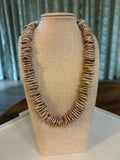 Beige necklace holder displaying a shell rondelle necklace. The holder is on a white plate and there is a green curtain in the background.