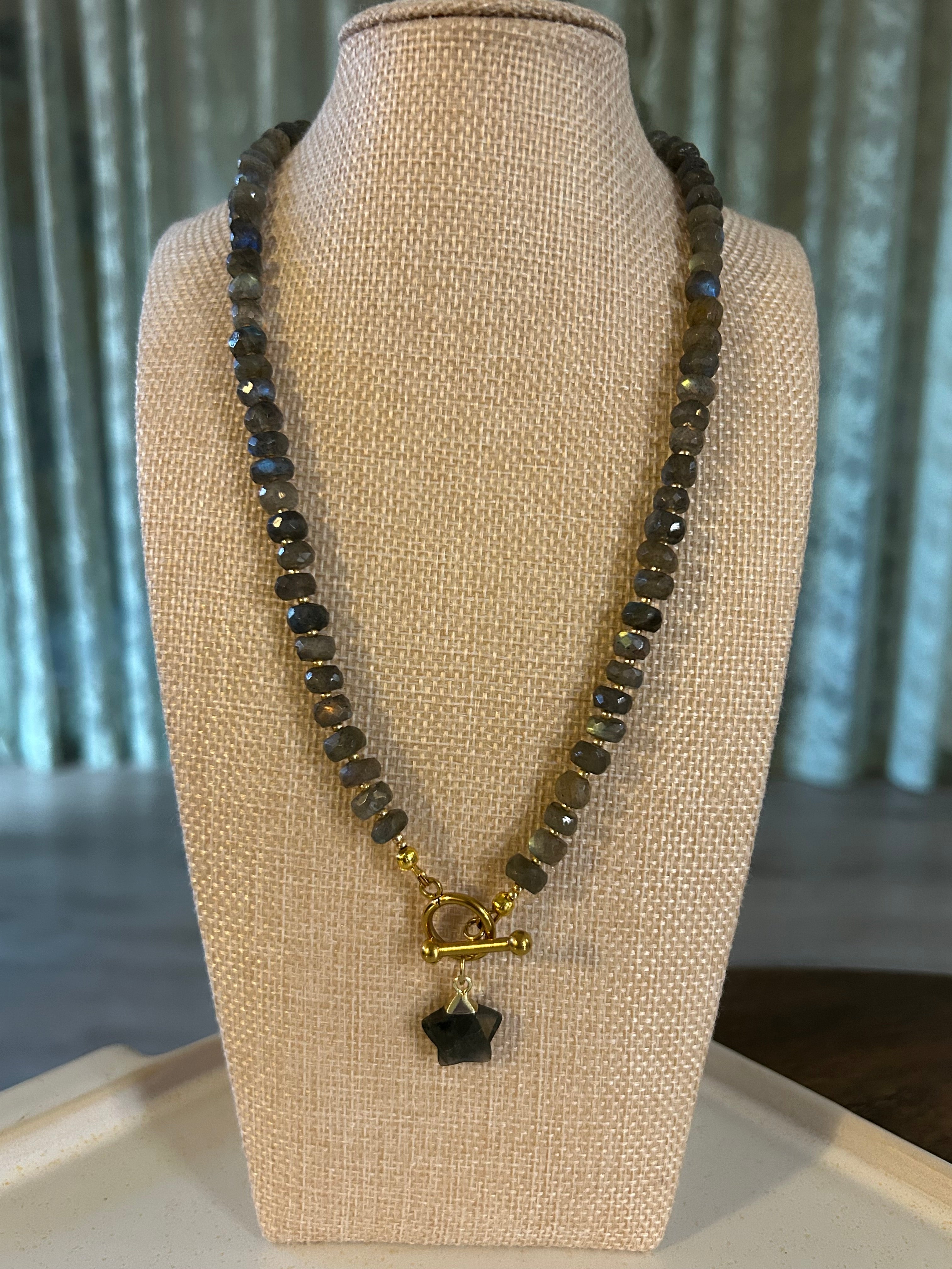 Necklace with gray beads and gold clasp on a beige display stand.